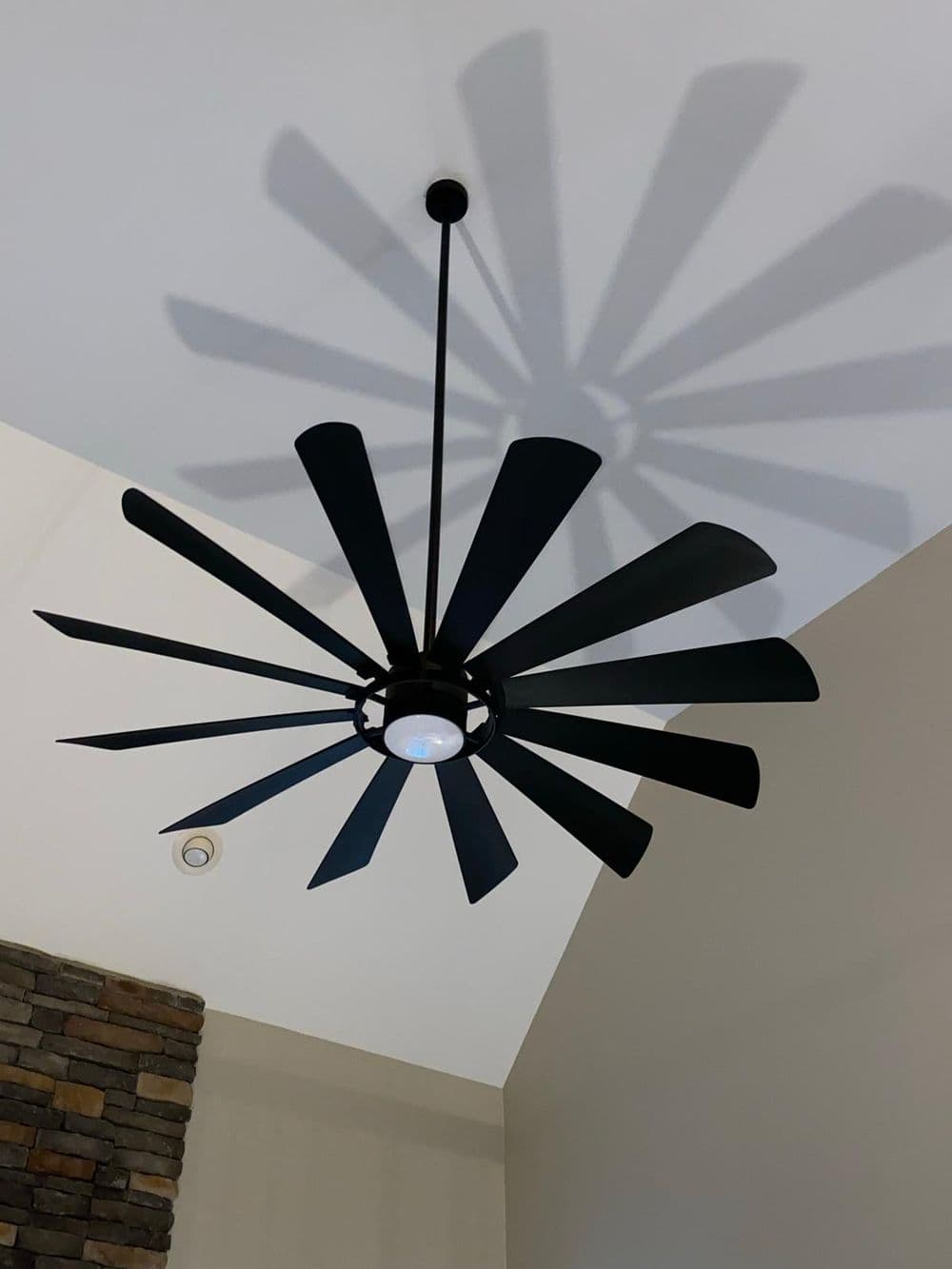 Black ceiling fan with wide blades casting a shadow on a beige wall and stone accent.