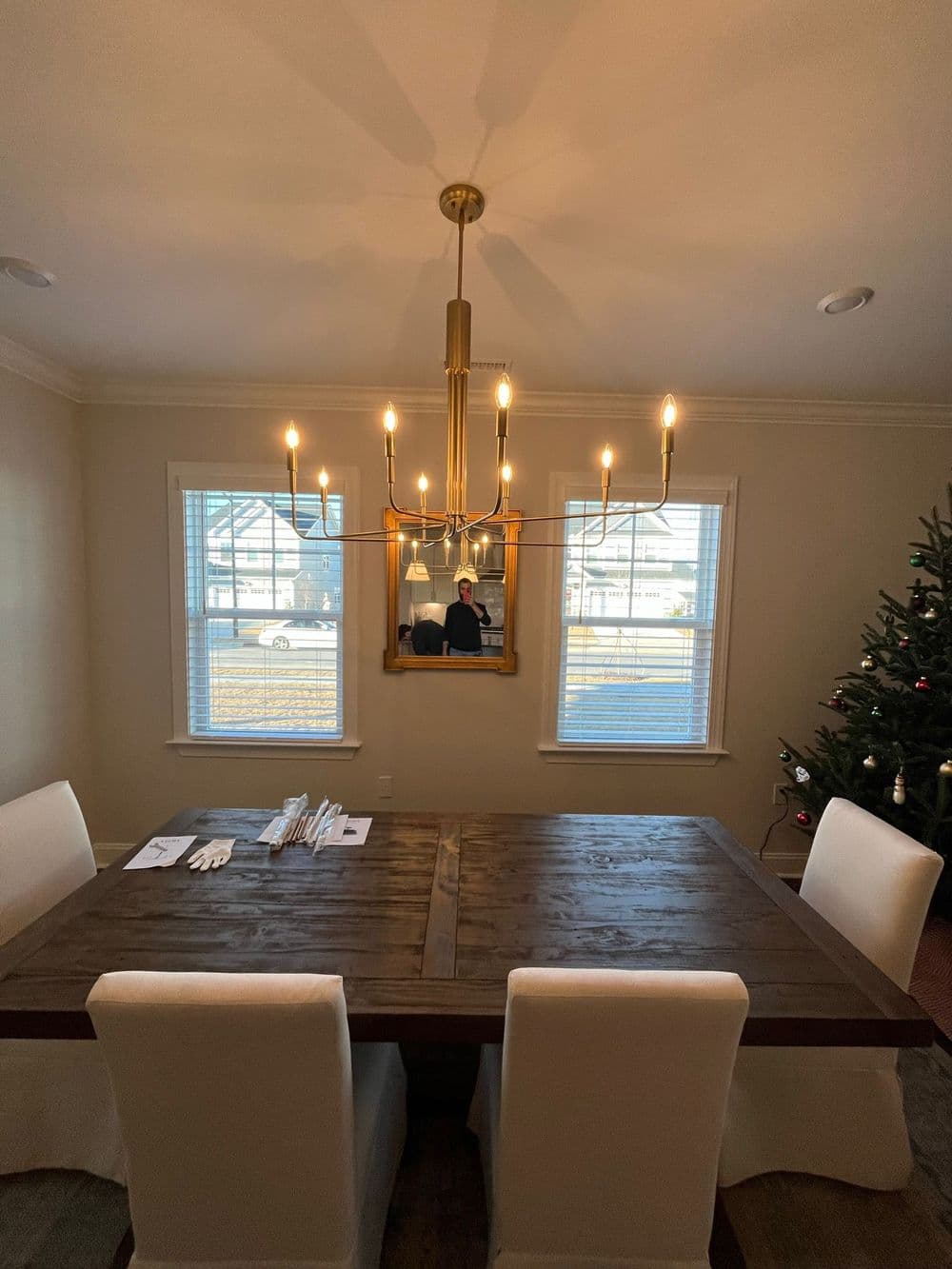 Modern dining room with wooden table, white chairs, chandelier, and Christmas tree in background.