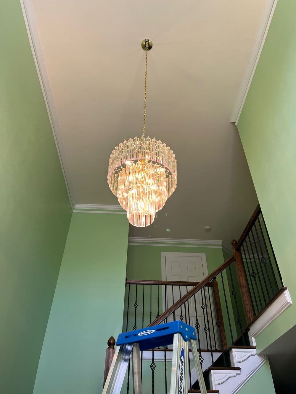 Elegant crystal chandelier hanging from a ceiling in a green-walled foyer with a ladder below.