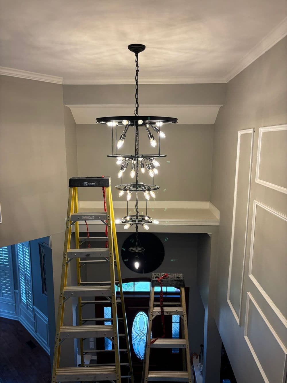 Modern chandelier installation with ladder in well-lit entryway. Elegant ceiling design.