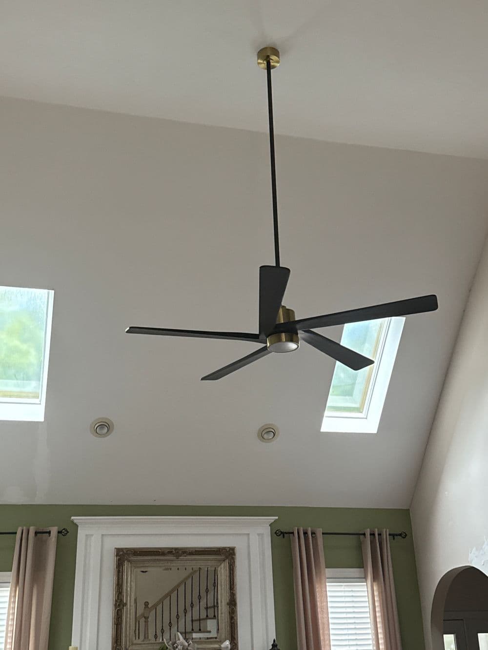 Ceiling fan with black blades and gold accents in a bright room with skylights.