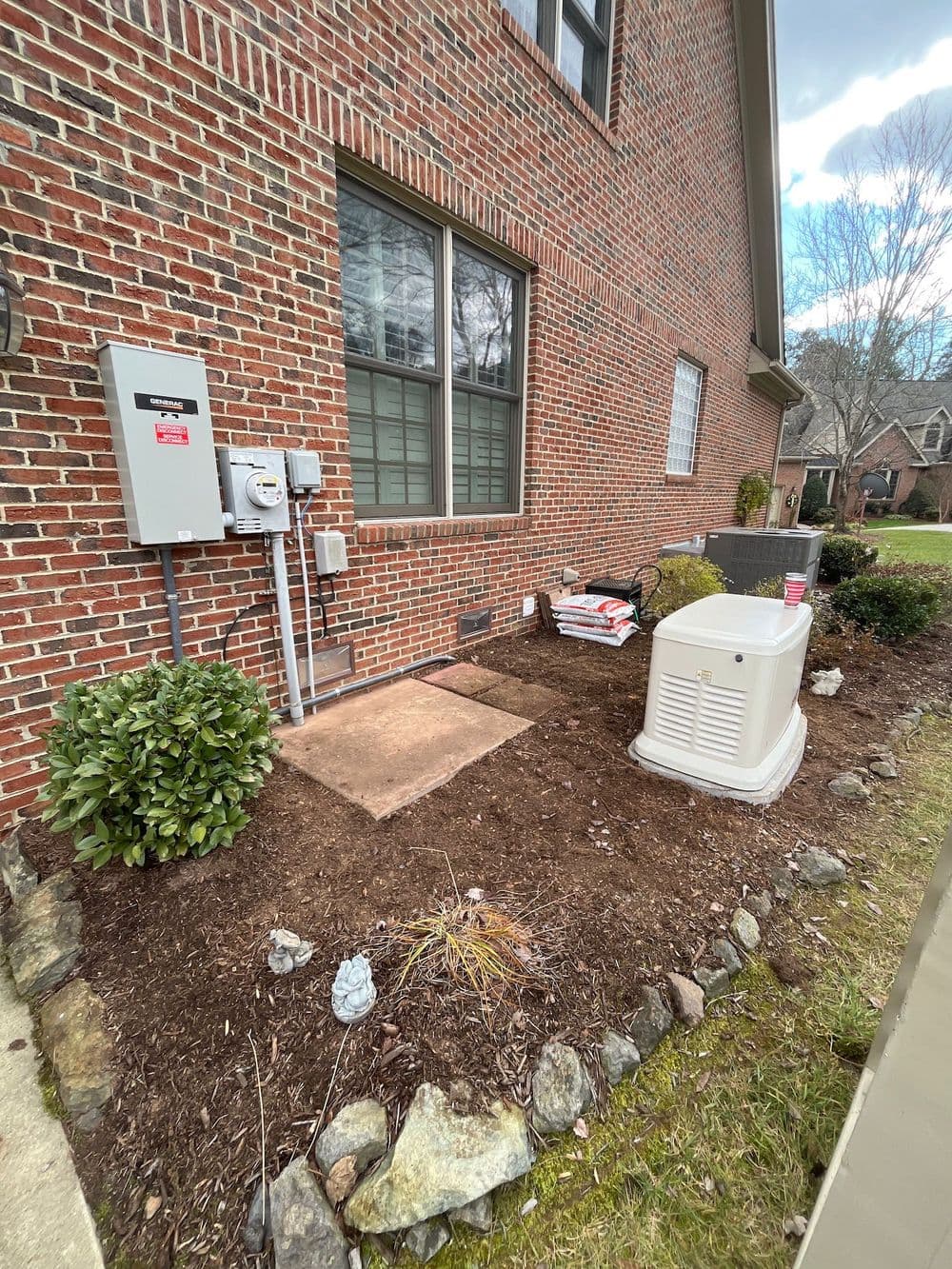 Garden area next to brick house with utility meters and outdoor equipment on gravel patch.
