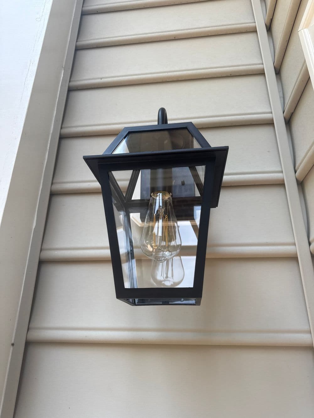 Black outdoor lantern with clear glass and hanging bulb mounted on beige siding.