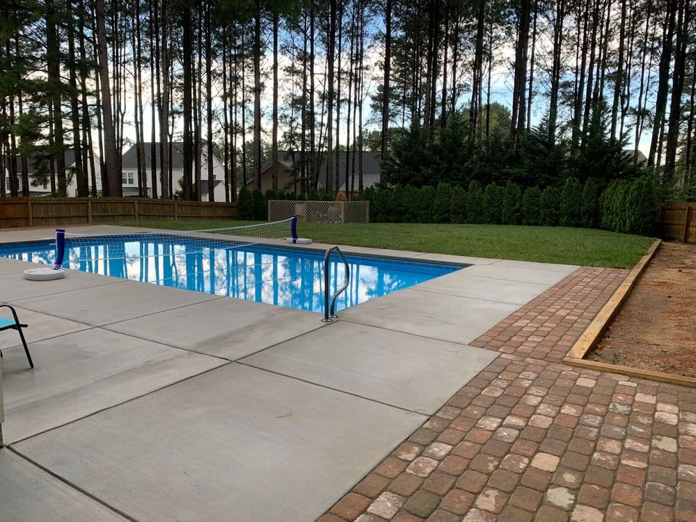 Backyard pool surrounded by lush trees and landscaped yard, featuring concrete and brick pathways.