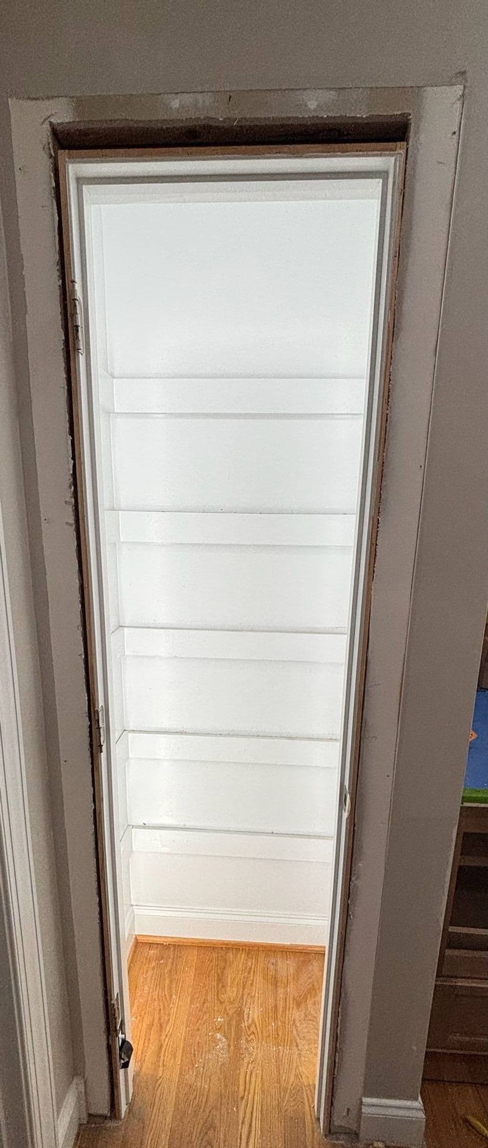 Empty white pantry with shelves, viewed from an open door, featuring wooden flooring.