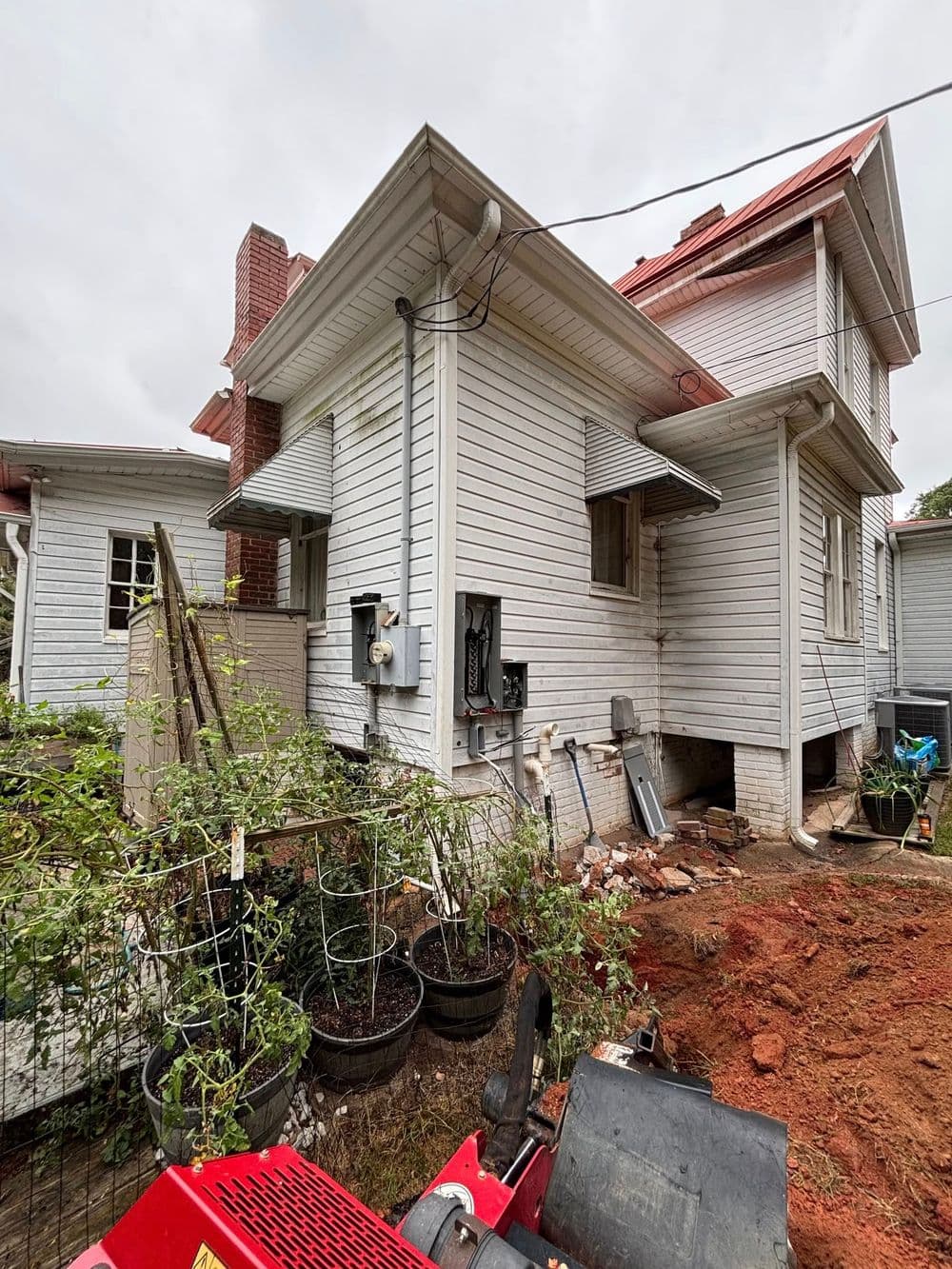 White two-story house with a red roof, overgrown garden, and gardening equipment in view.